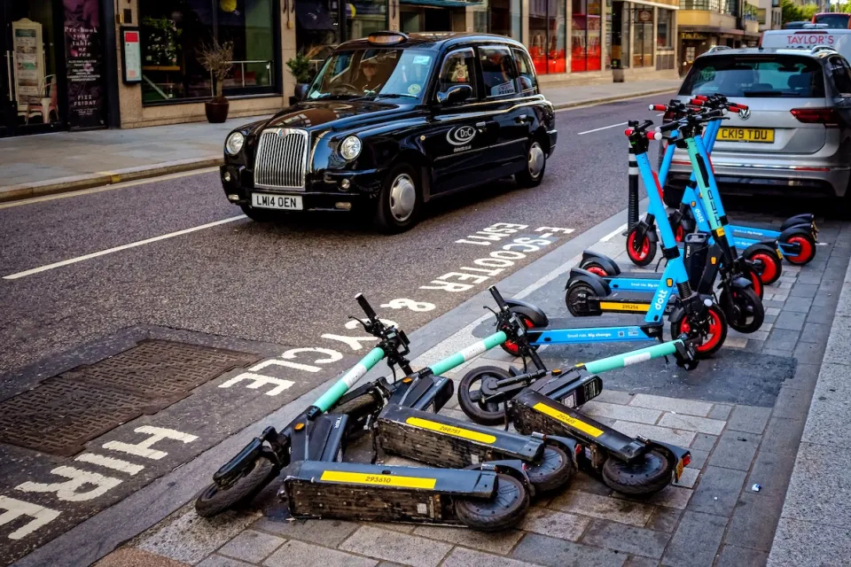 Group of e-scooters messily parked on street in London with black cab in background.